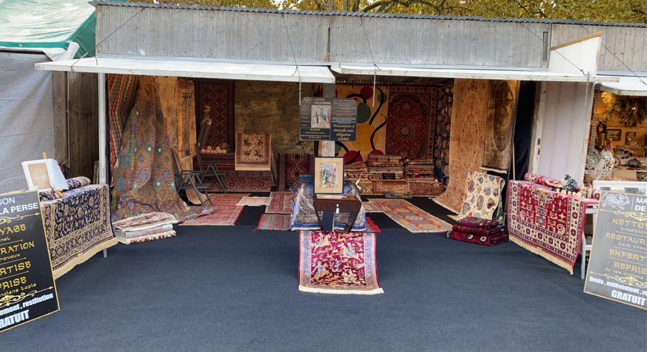 Le stand de La Maison de la Perse en plein air : tapis d'Orient suspendus et posés au sol, panneaux « Nettoyage, restauration, expertise, reprise »